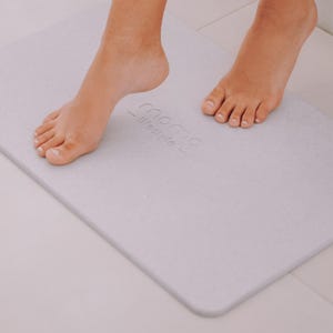 May include: A light gray bath mat with the text "momo - lifestyle -" printed on it. A person's feet are standing on the mat.