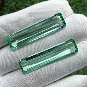 Pode incluir: Duas pedras preciosas alongadas de corte retangular com um tom verde-menta translúcido. Estas pedras facetadas apresentam bordas afiadas e limpas e uma superfície polida que reflete a luz, exibindo uma aparência e estrutura claras.