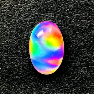 May include: An oval-shaped gemstone with a vibrant display of colors, including blue, green, yellow, orange, and pink. The polished surface reflects light, creating a shimmering effect. The gemstone is set against a textured black background.