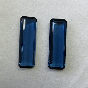 May include: Two rectangular, faceted blue gemstones with a deep navy hue. Each stone features a clear, polished surface with sharp, beveled edges and a long, slender profile, reflecting light across their smooth, transparent, and dark blue faces.