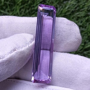Puede incluir: Una piedra preciosa de amatista larga, rectangular y facetada con un tono púrpura vibrante. La piedra presenta bordes afilados y limpios, y una superficie pulida y reflectante que capta la luz, mostrando su claridad transparente y corte.