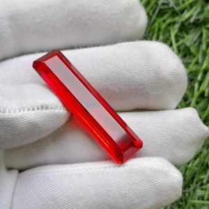 May include: A long, rectangular, faceted gemstone with a vibrant, translucent red colour. The stone features sharp, clean edges and a smooth, polished surface that reflects light, creating a bright, clear appearance across its elongated shape.