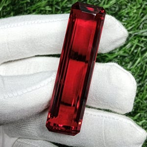 Puede incluir: Una piedra preciosa rectangular facetada de color rojo intenso y vibrante. La piedra presenta un corte tipo baguette largo y delgado con bordes afilados y limpios, y una superficie altamente pulida y reflectante con refracción de luz.