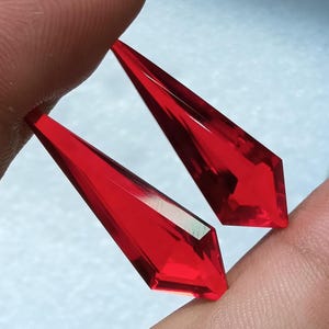 May include: Two elongated, pointed, kite-shaped red crystals with faceted surfaces. These translucent, vibrant red gemstones feature sharp, angular edges and a smooth, polished finish, reflecting light across their geometric, multi-faceted structure.