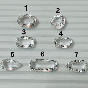 May include: Seven clear, faceted gemstones in various shapes: oval, triangular, and rectangular. The gemstones are arranged on a light gray surface. The facets reflect light, creating a sparkling effect. Ideal for jewelry making or collecting.