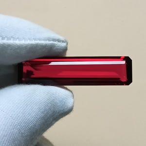 以下が含まれることがあります： 深く鮮やかな赤色の、長く長方形のファセットカットされた宝石。石は鋭くきれいなエッジと光を反射する磨かれた表面を持ち、その細長い形状全体に透明でクリアな品質を示しています。