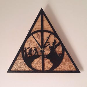 May include: Triangular wooden wall clock with a black frame and a light brown face. The clock features a laser-cut design depicting a scene with figures and a castle. The clock hands are black and the clock is mounted on a white wall.