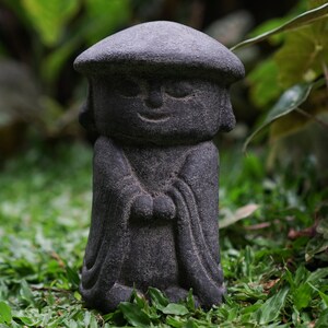 May include: A small, gray stone statue of a person wearing a hat and a robe. The statue is standing on a bed of green grass.