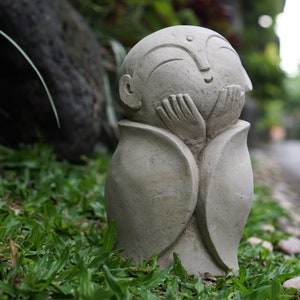 May include: A gray stone sculpture of a person with their hands clasped in front of their face. The sculpture is sitting on a bed of green grass.