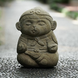 May include: A stone statue of a seated monkey with closed eyes, wearing a decorative headpiece and clothing. The sculpture is a light gray color and is set against a blurred outdoor background.