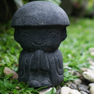 May include: A small, black stone statue of a person with a wide smile, wearing a hat and sitting with hands clasped in prayer.