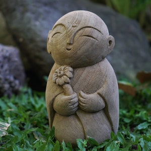 May include: A small, smiling stone statue of a person holding a flower. The statue is made of a light brown stone and has a simple, serene expression.