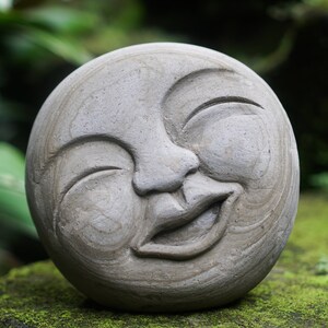 May include: A round, gray stone sculpture of a smiling face. The face has closed eyes, a wide, open mouth, and a slightly tilted head. The sculpture is set on a mossy surface, with green foliage in the background.