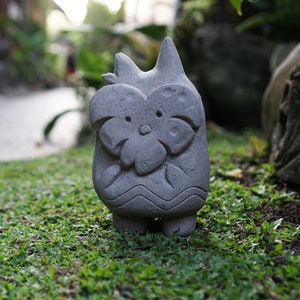 May include: A gray stone owl sculpture with a flower-like face and a wavy pattern at the base. The owl has large eyes and a pointed head. It stands on green grass in a garden setting.