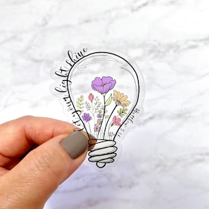 May include: Clear sticker featuring a lightbulb design filled with colorful flowers. The words "Let your light shine" are written around the bulb, with "Matthew 5:16" in the corner. The flowers include purple, pink, and yellow blooms.