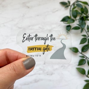 May include: Clear sticker with a yellow and gray design. The sticker features a road leading to a sunrise with the text "Enter through the narrow gate" and the scripture reference "Matthew 7:13-14".