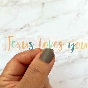May include: A clear sticker with the words "Jesus loves you" in pastel colors. The sticker is held by a hand with a neutral-toned nail polish. The text is in a handwritten style.