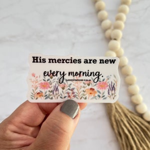 May include: A clear sticker with the text "His mercies are new every morning" and the Bible verse Lamentations 3:22-23. The sticker features a floral design with pink, orange, and purple flowers. The sticker is held by a hand.