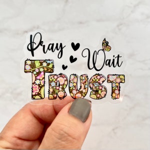 May include: A clear sticker with the words "Pray Wait Trust" in black text. The word "Trust" is decorated with a floral pattern in brown, pink, and yellow.