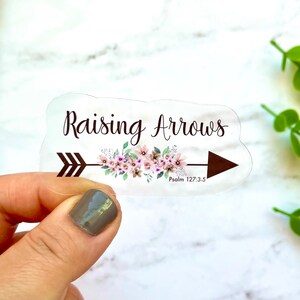 May include: Clear sticker with a brown arrow and pink floral design. The text "Raising Arrows" is above the arrow and the text "Psalm 127:3-5" is below the arrow.