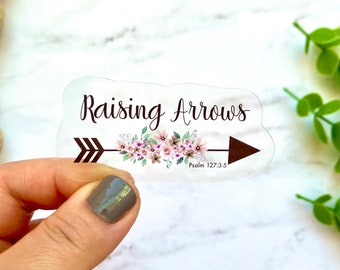 May include: Clear sticker with a brown arrow and pink floral design. The text "Raising Arrows" is above the arrow and the text "Psalm 127:3-5" is below the arrow.