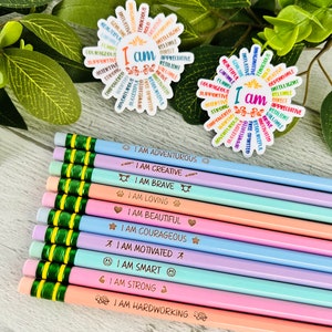 Puede incluir: L&aacute;pices de colores pastel con frases motivadoras como "Soy valiente" y "Soy fuerte". Tambi&eacute;n se ven dos pegatinas con forma de flor con las palabras "Yo soy" rodeadas de afirmaciones positivas.