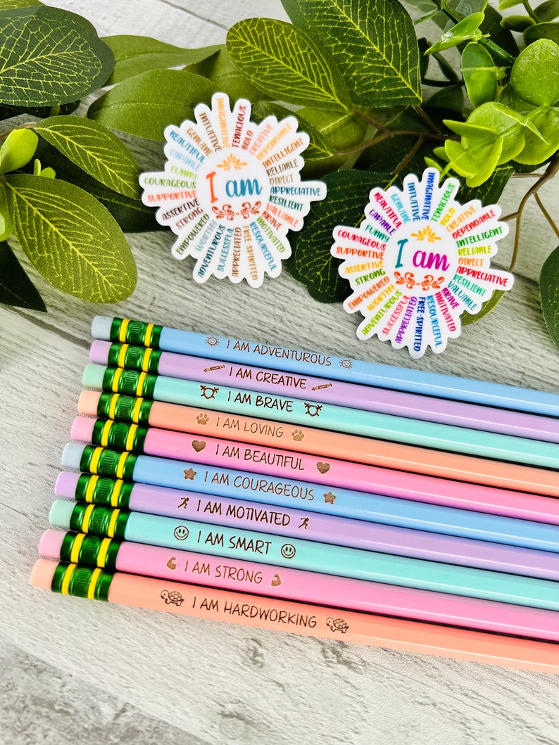 Puede incluir: L&aacute;pices de colores pastel con frases motivadoras como "Soy valiente" y "Soy fuerte". Tambi&eacute;n se ven dos pegatinas con forma de flor con las palabras "Yo soy" rodeadas de afirmaciones positivas.