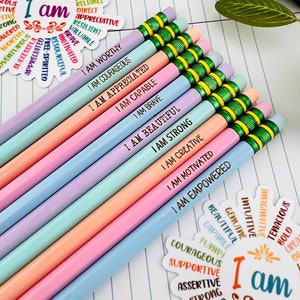 Puede incluir: Un conjunto de l&aacute;pices de colores pastel con frases motivadoras como "I AM WORTHY" y "I AM COURAGEOUS" impresas. Los l&aacute;pices est&aacute;n colocados sobre un cuaderno rayado, acompa&ntilde;ados de pegatinas decorativas con afirmaciones positivas.