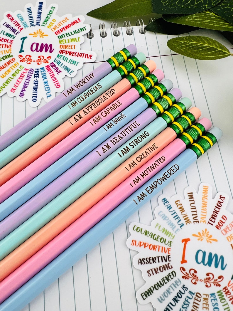 Puede incluir: Un conjunto de l&aacute;pices de colores pastel con frases motivadoras como "I AM WORTHY" y "I AM COURAGEOUS" impresas. Los l&aacute;pices est&aacute;n colocados sobre un cuaderno rayado, acompa&ntilde;ados de pegatinas decorativas con afirmaciones positivas.