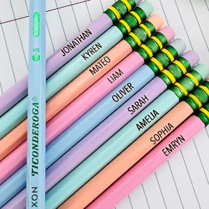 Puede incluir: Una colecci&oacute;n de l&aacute;pices de colores pastel, cada uno con un nombre impreso en el lateral. Los l&aacute;pices est&aacute;n dispuestos en diagonal sobre un cuaderno rayado. Un l&aacute;piz es de color azul claro con el texto "Dixon Ticonderoga". Los l&aacute;pices tienen detalles verdes y amarillos.