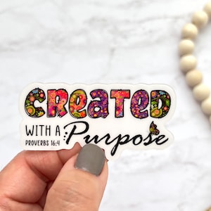 May include: A white sticker with the words "Created with a Purpose" in black text. The word "Created" is in colorful patterned letters. The sticker also includes the text "Proverbs 16:4" and a small butterfly graphic.