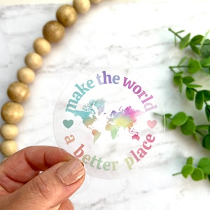 May include: A clear sticker with a rainbow-colored world map and the text "make the world a better place" with hearts on either side of the text.