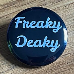 May include: A circular pin-back button with a glossy dark navy blue background. The text "Freaky Deaky" is printed in a light blue, sans-serif font across the centre of the button, with "Freaky" on the top line and "Deaky" on the bottom line.