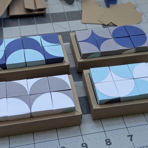 May include: Four sets of square blocks arranged in rectangular cardboard trays. Each set features geometric patterns in shades of blue, gray, and white, including semi-circles, circles, and star shapes printed on the top surfaces of the blocks.