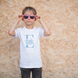 May include: A white t-shirt with a blue letter E and the name "Esther" written inside the letter. A young person is wearing the shirt and pink sunglasses.
