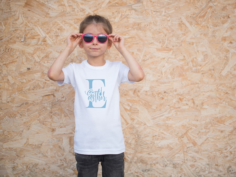 May include: A white t-shirt with a blue letter E and the name "Esther" written inside the letter. A young person is wearing the shirt and pink sunglasses.