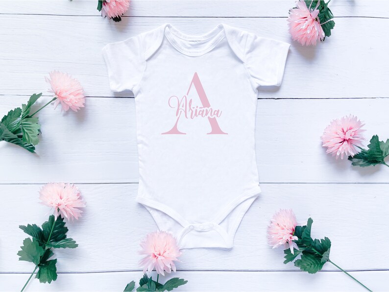 May include: White baby onesie with a pink letter A and the name "Ariana" written in pink cursive.