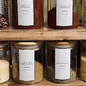 Puede incluir: Frascos de vidrio llenos de diversas hierbas y especias, incluyendo chile en polvo, chipotle en polvo, cilantro molido y mejorana. Cada frasco tiene una etiqueta blanca con el nombre de la especia en negro. Los frascos están colocados en estantes de madera.