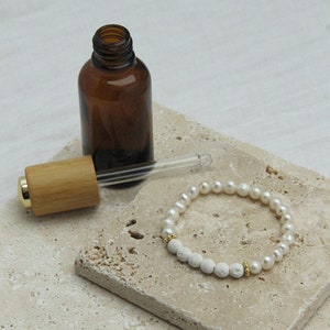 Könnte beinhalten: Eine braune Glasflasche mit einem Bambus-Tropfer und einem weißen Perlen- und Lavastein-Armband auf einer beige gefliesten Oberfläche.