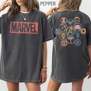 Puede incluir: Camiseta gris oscuro con la palabra "MARVEL" en un rectángulo rojo en la parte delantera. La parte trasera de la camiseta presenta un diseño hexagonal colorido con varios símbolos de superhéroes, incluido el logotipo de los Vengadores.