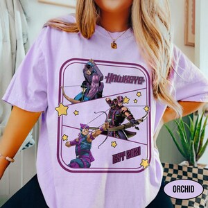 Puede incluir: Camiseta lila claro con un gr&aacute;fico del superh&eacute;roe Hawkeye. El dise&ntilde;o incluye ilustraciones de Hawkeye, la palabra "Hawkeye" y el texto "EST 2021". La camiseta tiene cuello redondo y mangas cortas.