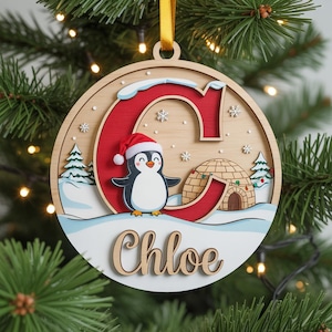 May include: A wooden Christmas ornament featuring a penguin wearing a Santa hat, snowflakes, and an igloo. The letter "C" is in red, and the name "Chloe" is written below. The ornament is round with a gold ribbon for hanging.