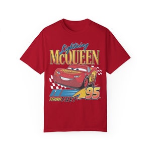 Pu&ograve; includere: Maglietta rossa con una grafica di Saetta McQueen dal film Cars della Disney Pixar. La grafica include il testo "Lightning McQueen" e "Think Fast" con una bandiera a scacchi e il numero 95.