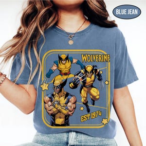 Puede incluir: Camiseta azul vaquero con un gr&aacute;fico de Wolverine. El gr&aacute;fico incluye tres ilustraciones de Wolverine en amarillo y azul, con la palabra "Wolverine" y "EST 1974" en texto amarillo. La camiseta tiene cuello redondo.