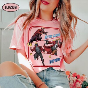 Puede incluir: Camiseta rosa claro con un gr&aacute;fico de Star Lord. El gr&aacute;fico incluye ilustraciones de Star Lord en poses de acci&oacute;n, las palabras "STAR LORD" y "EST 2014" en un estilo retro. La camiseta tiene cuello redondo.