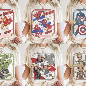 Puede incluir: Camisetas color crema con dise&ntilde;os de superh&eacute;roes de Marvel. Cada camiseta muestra un personaje o equipo diferente, incluyendo Iron Man, Spider-Man, Capit&aacute;n Am&eacute;rica, Hulk, Thor y Groot, con el nombre del personaje y el a&ntilde;o de establecimiento.