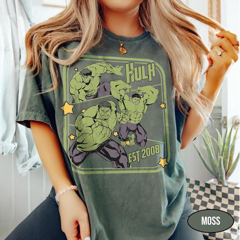 Puede incluir: Camiseta verde musgo con un dise&ntilde;o gr&aacute;fico de Hulk en varias poses, la palabra "Hulk" y "EST 2008" en un marco verde, y estrellas amarillas. La camiseta es de estilo casual.