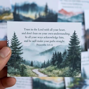 May include: A rectangular card featuring a watercolor landscape with a path winding through a forest. The text "Trust in the Lord... Proverbs 3:5-6" is printed on the card. The card is held in a person's hand.