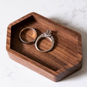 May include: A wooden ring dish with a hexagonal shape, made of dark walnut wood. Two silver rings are resting inside the dish, one with a diamond.