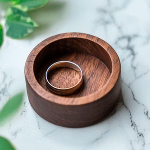 May include: A wooden ring dish with a dark brown finish. A silver ring is resting inside the dish.
